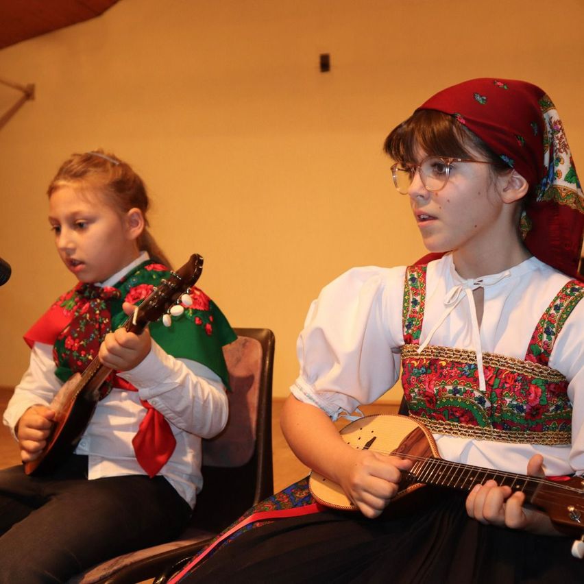 Zwei Mädchen in traditioneller Kleidung spielen Saiteninstrumente. Eine sitzt auf einem Stuhl, die andere steht. Sie befinden sich in einem Raum mit gelber Wand.