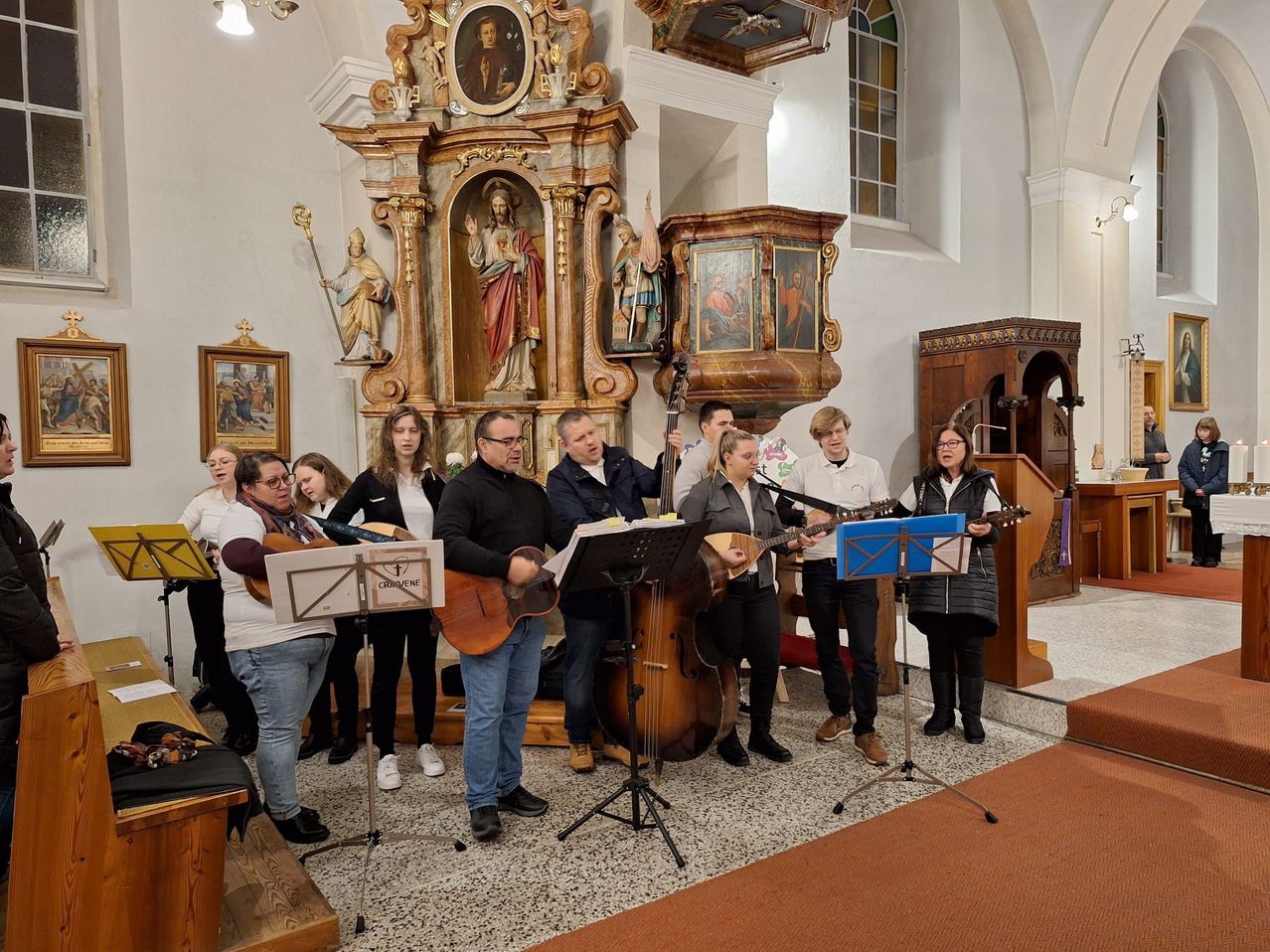 Eine Gruppe von Menschen steht in einer Kirche und spielt Musikinstrumente. Sie stehen vor einem Podium und halten Gitarren, eine Geige und eine Mandoline. Hinter ihnen steht ein hölzerner Altar mit einer Statue von Jesus Christus. Auf der linken Seite steht eine Statue eines Heiligen.