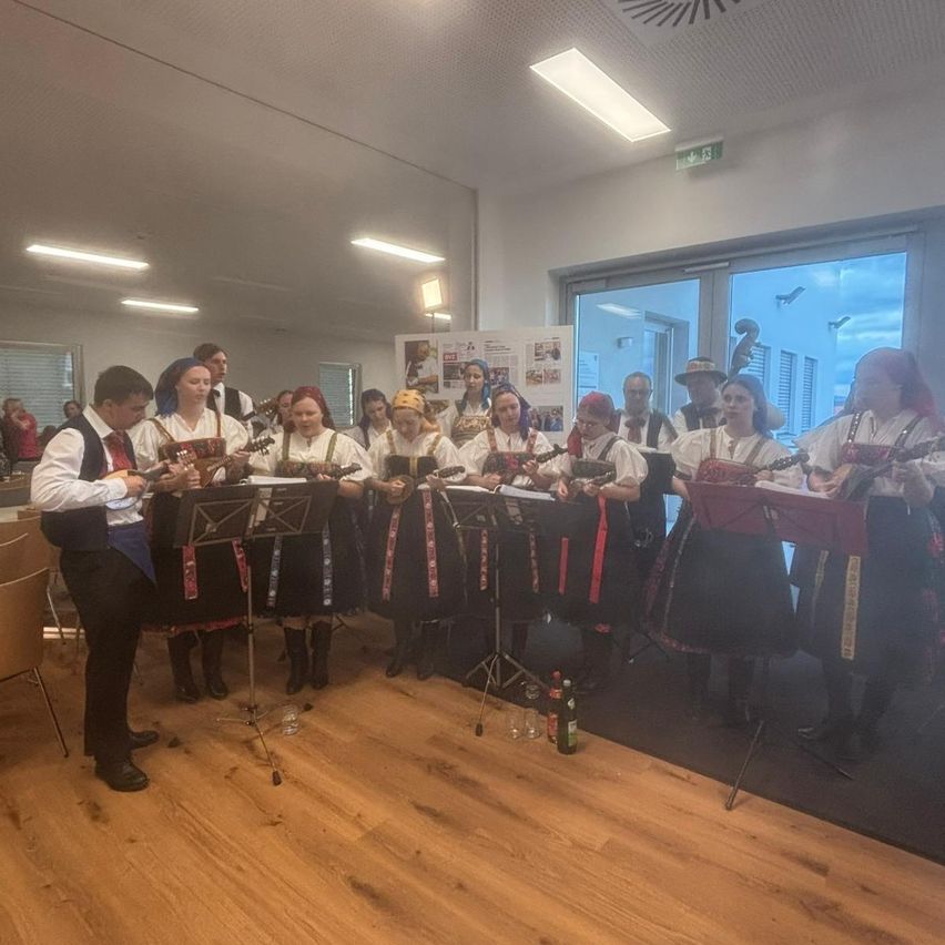Eine Gruppe von Musikern in traditioneller Kleidung tritt auf der Bühne auf. Sie spielen Instrumente, während ein Mann vorne steht. Der Raum hat Holzböden, Stühle und eine Glastür. Es gibt auch Lampen an der Decke.