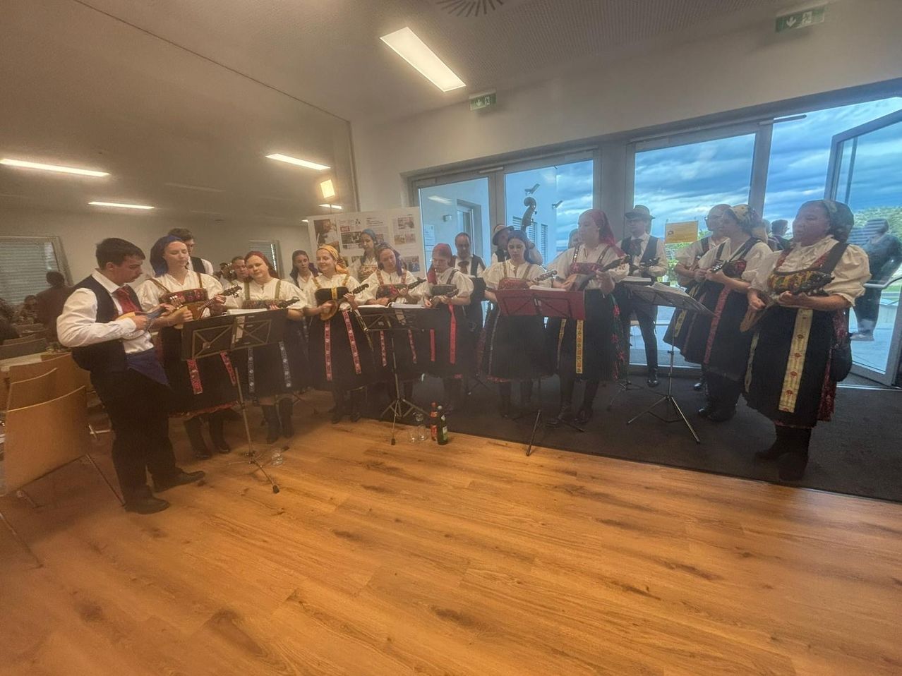 Eine Gruppe von Musikern in traditioneller Kleidung steht in einem Raum mit Holzböden und spielt Instrumente vor einem Glasfenster.