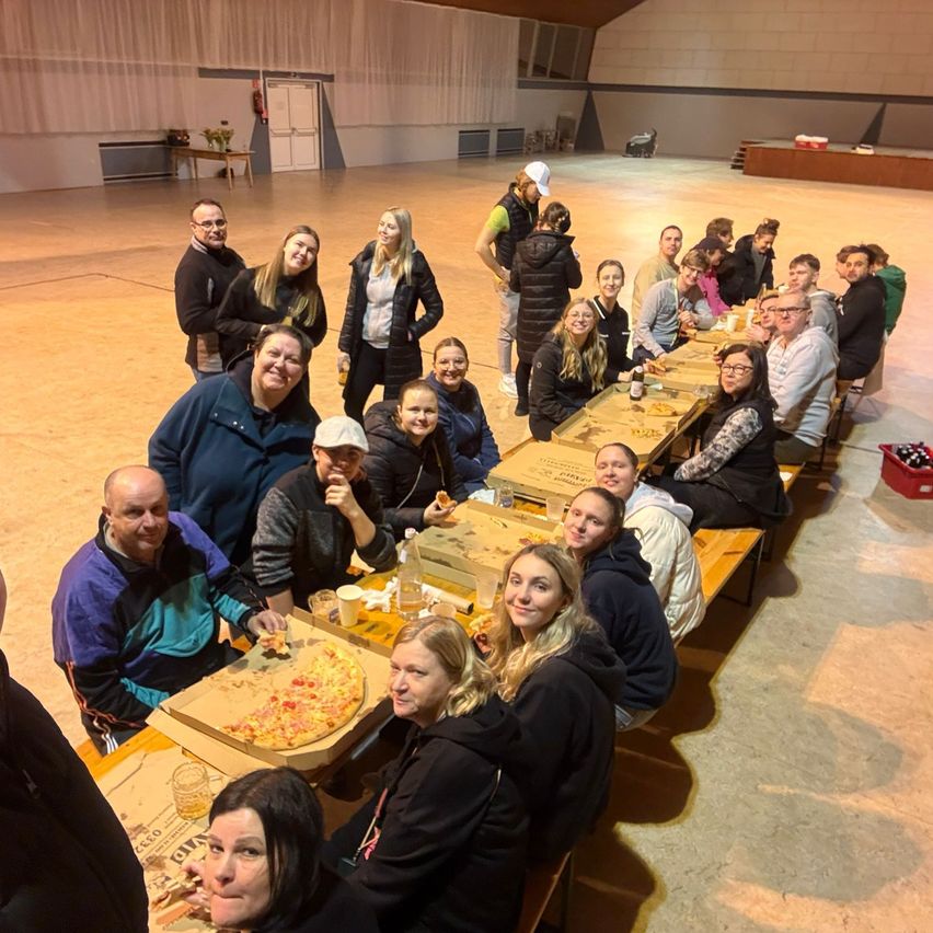A group of people are gathered around long tables in a large room, enjoying pizza. Some are seated while others stand behind the tables. A woman holds a pizza slice.