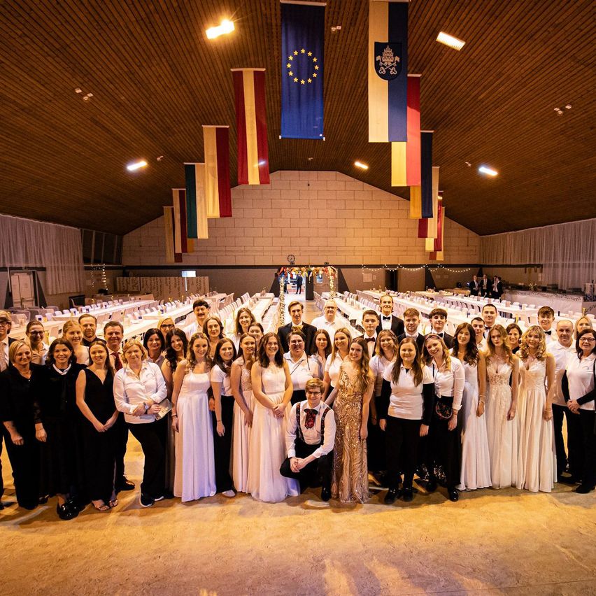 A large group of people dressed in formal attire are posing for a group photo in a banquet hall. Flags and lights decorate the space.