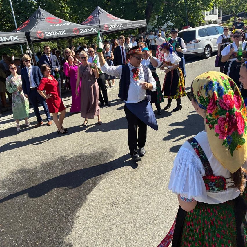 A man in a traditional costume holds a bottle of beer while dancing in the street, surrounded by people in formal attire and traditional costumes.