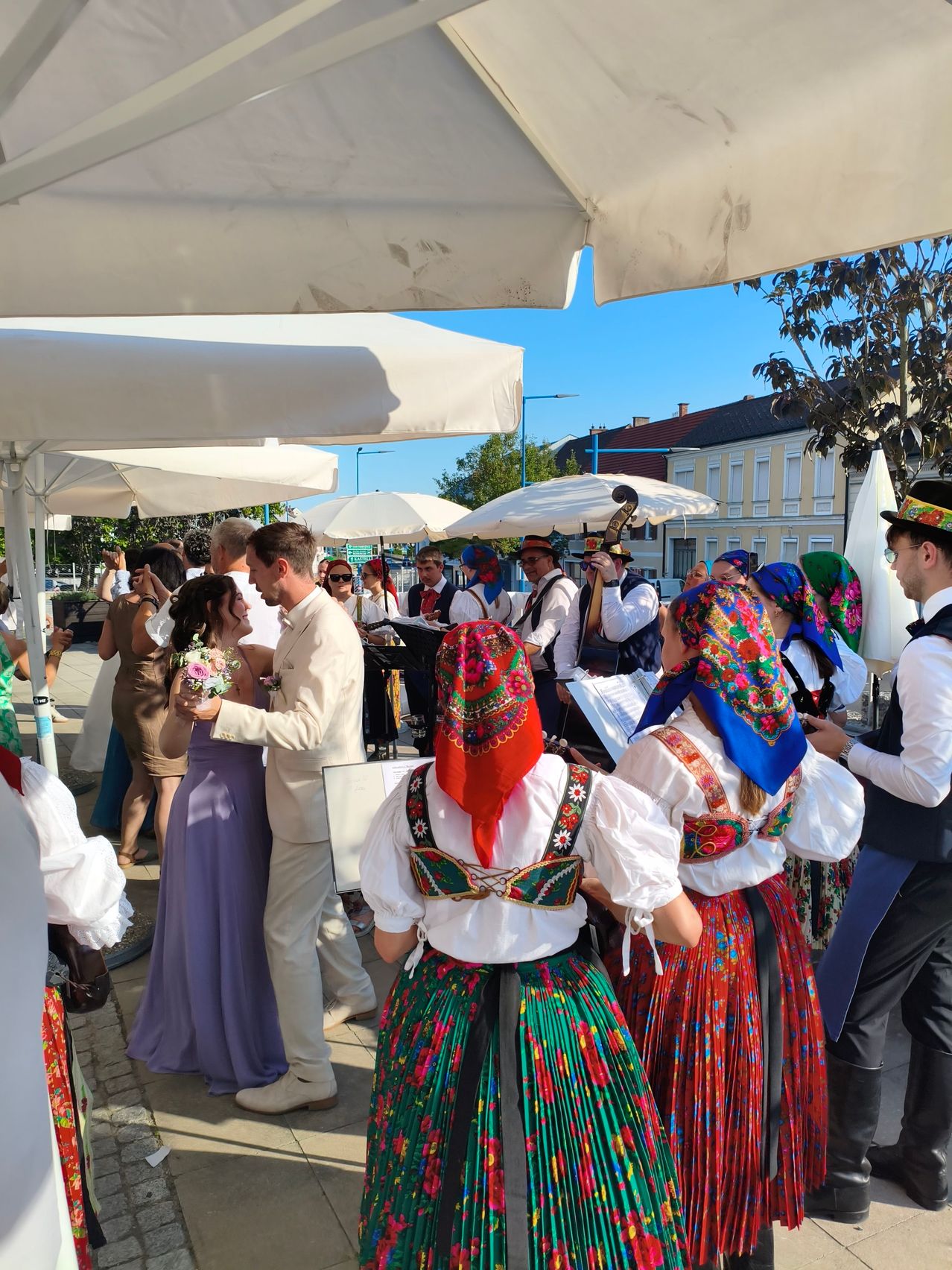 A wedding ceremony outdoors with a band playing, the bride and groom in the foreground, and several people in traditional costumes.