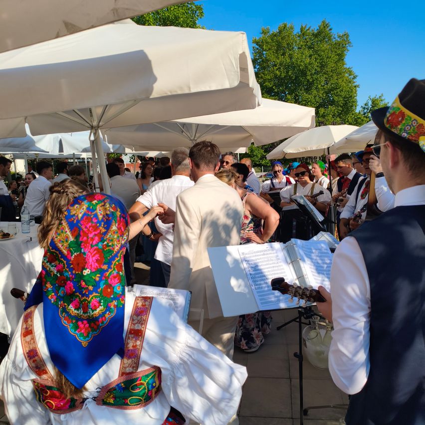 A group of people in traditional attire are dancing and enjoying a performance by a band under white umbrellas in an outdoor setting.