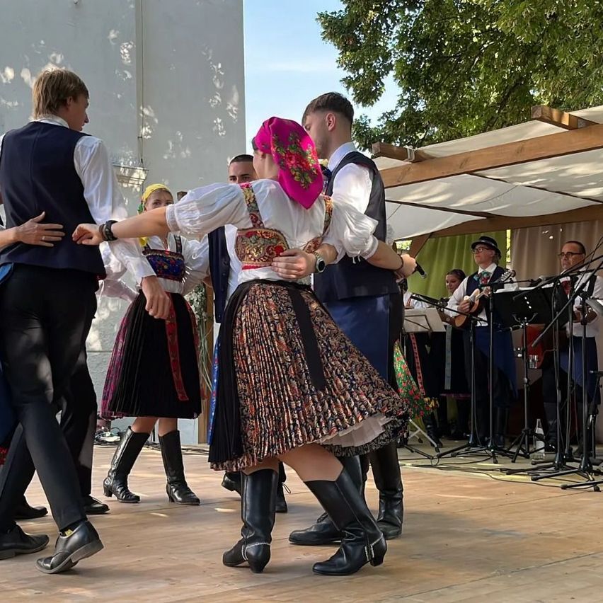 A group of people dressed in traditional folk costumes are dancing on a stage with a band playing behind them.