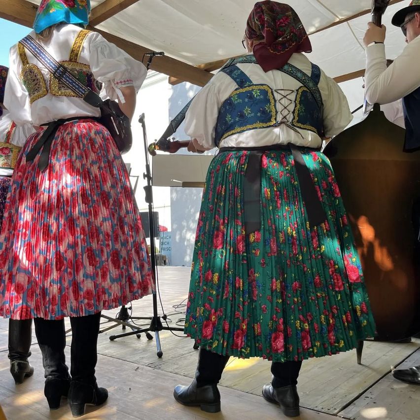 Three people in traditional folk costumes stand on a stage with musical instruments. The person in the middle plays a cello, while the others are holding guitars.