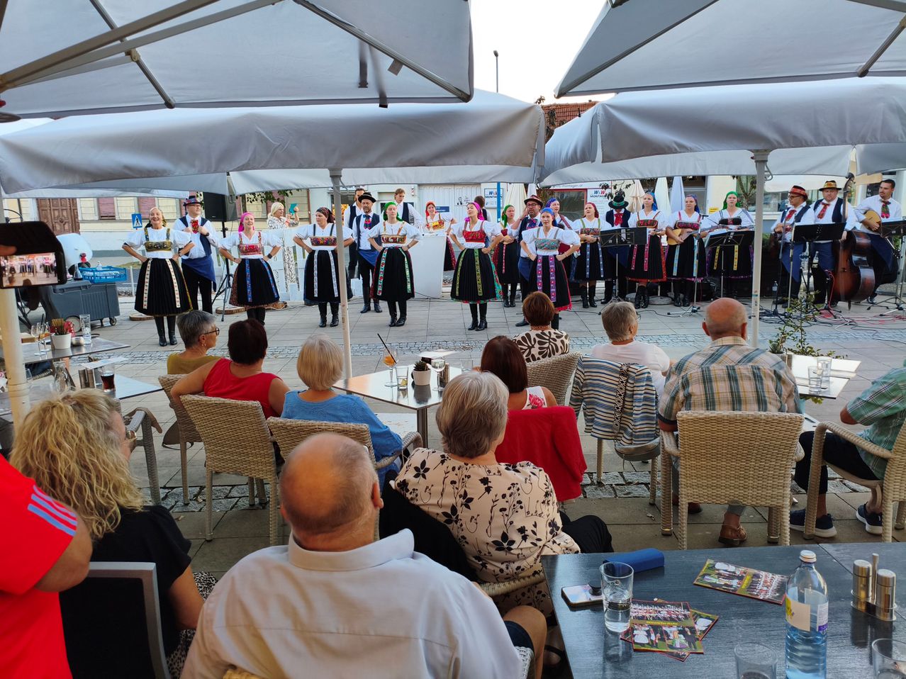 A group of people dressed in traditional costumes perform on a stage in an outdoor setting, with an audience seated in chairs under umbrellas.