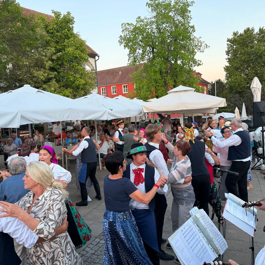 A group of people are dancing in an outdoor area with white umbrellas. Some are wearing traditional clothing. There are trees and buildings in the background.