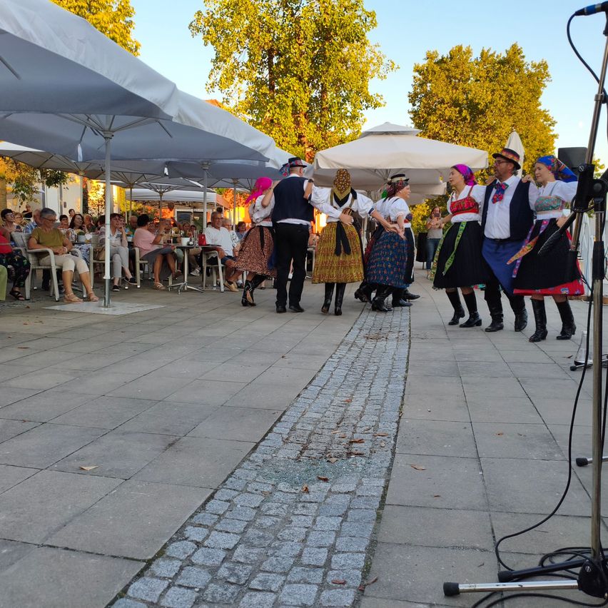 A group of people in traditional costumes are performing a dance on a paved area with umbrellas and a microphone. Spectators are seated under the umbrellas watching.