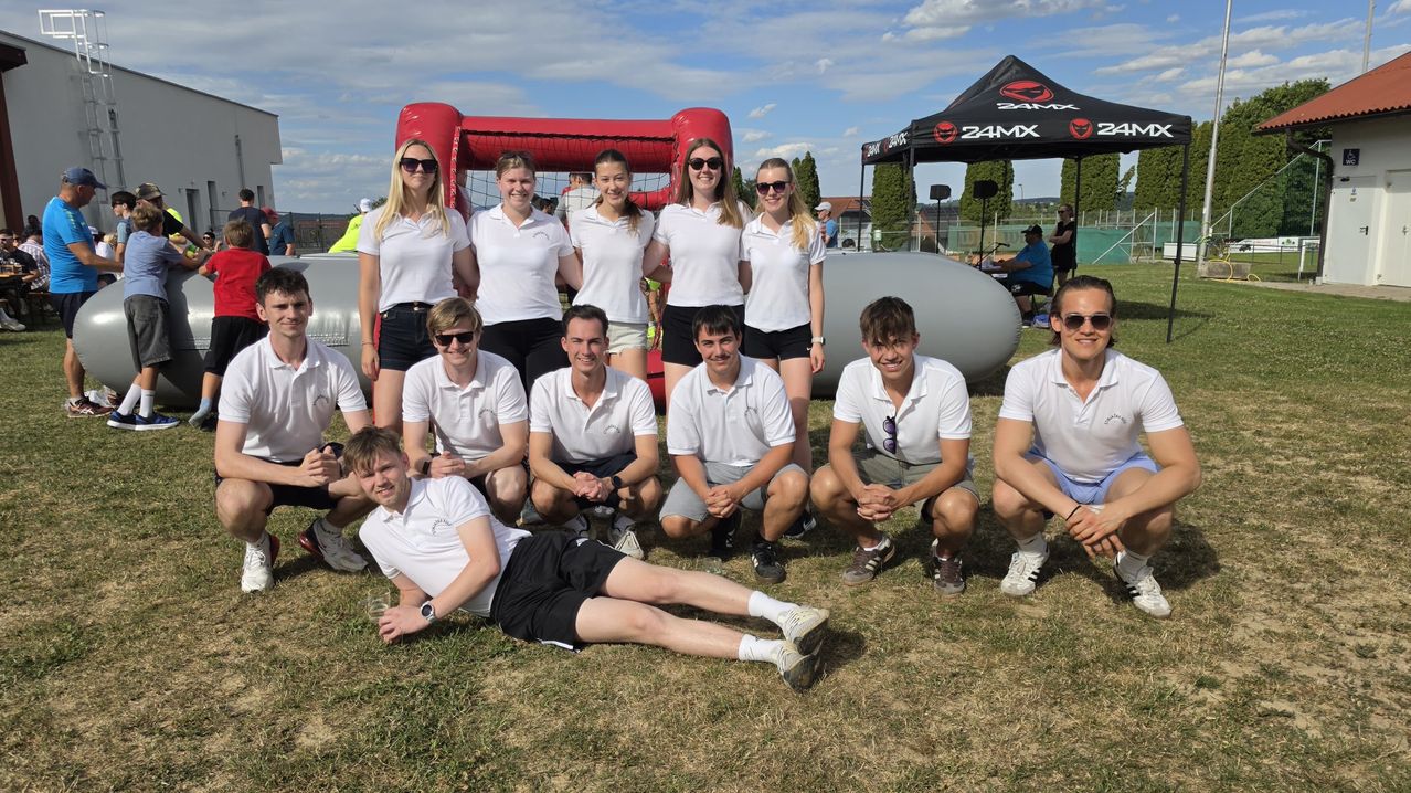 A group of people in white shirts and black shorts pose for a photo on a grassy field with an inflatable obstacle in the background.
