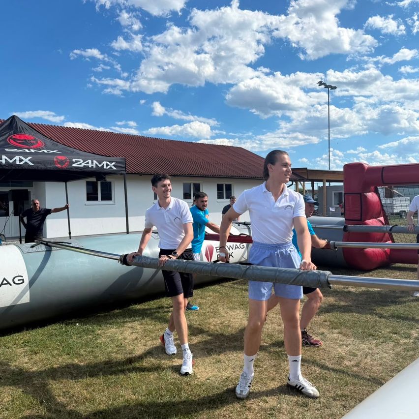 Three individuals are participating in an outdoor event, holding a long metal pole. The scene takes place in front of a white building with a red roof and a tent with the text 24MX.