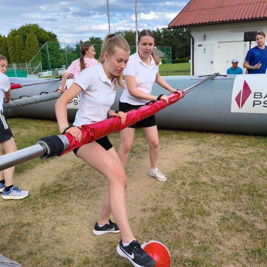 A group of young women are engaging in an outdoor activity. They are holding a long red pole, likely participating in a game. They are dressed in white polo shirts and black shorts. Nearby, an inflatable obstacle and a building with a red roof are visible.