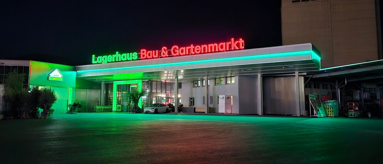 Ein Gebäude mit einem roten Schild, auf dem 'Lagerhaus Bau & Gartenmarkt' steht. Das Gebäude ist mit grünen Lichtern beleuchtet. Ein Auto ist vor dem Gebäude geparkt.