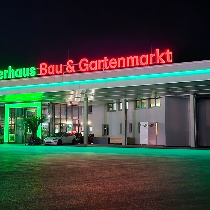 Ein Gebäude mit einem roten Schild, auf dem 'Lagerhaus Bau & Gartenmarkt' steht. Das Gebäude ist mit grünen Lichtern beleuchtet. Ein Auto ist vor dem Gebäude geparkt.