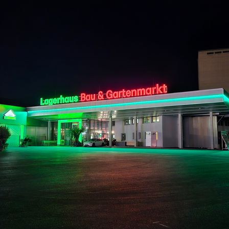 Ein großes Gebäude mit einem weißen Schild, auf dem Lagerhaus Bau & Gartenmarkt steht. Das Gebäude ist mit grünen Lichtern beleuchtet, und ein Auto ist davor geparkt.