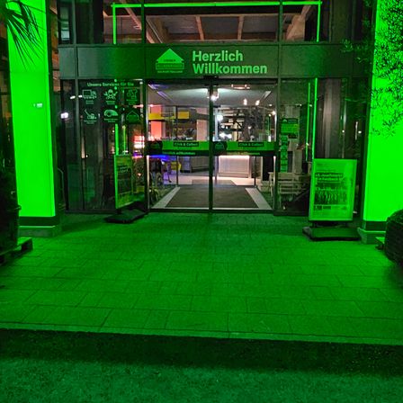 Der Eingang eines Gebäudes ist mit grünen Lichtern beleuchtet. Es steht Herzlich Willkommen (Willkommen) in weißen Buchstaben. Es gibt zwei Glastüren und einen mit Teppich ausgelegten Weg, der zum Eingang führt.