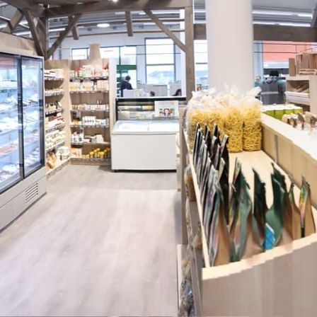 Ein Innenansicht eines Geschäfts mit Kühlvitrinen, Holzregalen und verschiedenen Lebensmitteln. Im Hintergrund sind zwei Personen zu sehen.