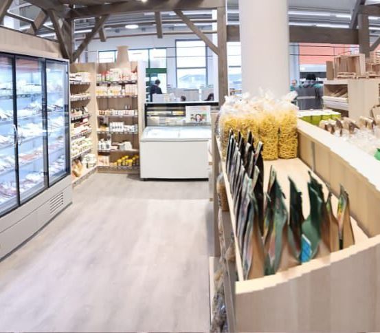 Ein Innenansicht eines Geschäfts mit Kühlvitrinen, Holzregalen und verschiedenen Lebensmitteln. Im Hintergrund sind zwei Personen zu sehen.