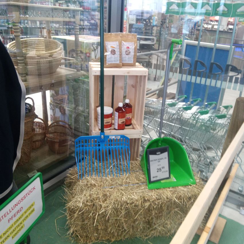 A hay bale with a blue pitchfork and a green dustpan is in a store display window. Behind it, there is a wooden crate with jars and a basket. Shopping carts are visible in the background.