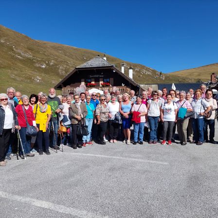 Eine Gruppe älterer Menschen steht vor einem Bergchalet. Einige tragen Brillen und halten Gehstöcke. Dahinter steht ein Haus mit roten Blumen und einem Schild, das Zechner Alm steht.