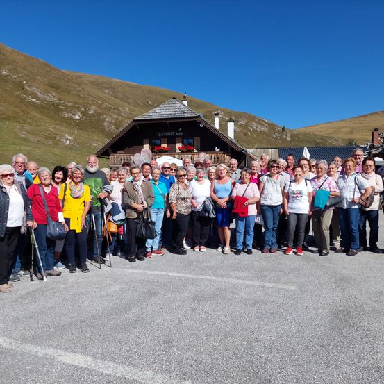 Eine Gruppe älterer Menschen steht vor einem Bergchalet. Einige tragen Brillen und halten Gehstöcke. Dahinter steht ein Haus mit roten Blumen und einem Schild, das Zechner Alm steht.