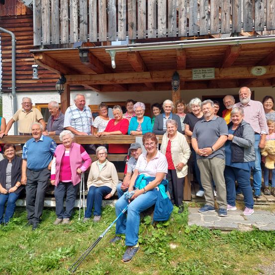 Eine Gruppe von Menschen steht und sitzt vor einem Holzhaus, lächelt und posiert für ein Foto. Einige haben Stöcke, Brillen und Hüte. Ein Schild befindet sich über dem Eingang.