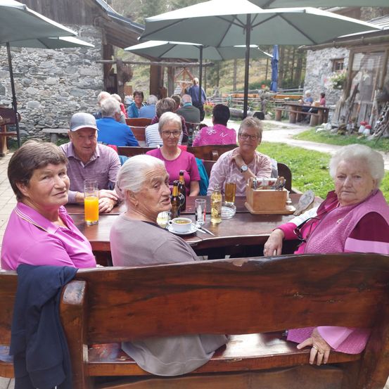 Eine Gruppe älterer Frauen und Männer sitzt um einen Tisch in einem Outdoor-Restaurant. Sie genießen Getränke und Snacks. Im Hintergrund befinden sich Schirme und eine Steinmauer.