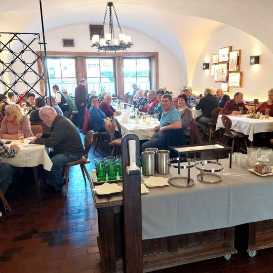 Ein Restaurant mit gewölbten Decken und Holzboden, mit mehreren Tischen und Stühlen. Menschen sitzen und unterhalten sich. Ein Tisch ist mit Essen beladen, und ein Tresen hat grüne Gläser und eine Tasse.