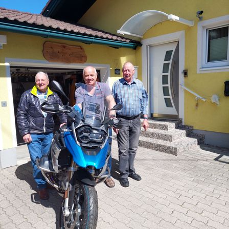 Drei Männer stehen vor einem gelben Haus mit einem blauen Motorrad. Ein Mann hält das Lenkrad, und ein anderer hat eine Uhr. Das Haus hat eine weiße Tür, ein Fenster und eine Garage.