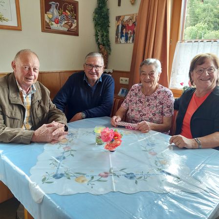 Vier Menschen sitzen um einen Tisch herum. In der Mitte sitzt eine Frau mit Blumen vor ihr. Auf der linken Seite ist ein Mann in einer Jacke. Hinter ihnen ist eine Wand mit einem Bilderrahmen und einer Pflanze.