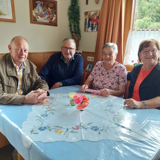Vier Menschen sitzen um einen Tisch herum. In der Mitte sitzt eine Frau mit Blumen vor ihr. Auf der linken Seite ist ein Mann in einer Jacke. Hinter ihnen ist eine Wand mit einem Bilderrahmen und einer Pflanze.