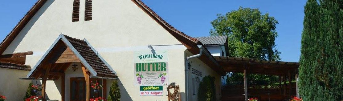 Ein traditionelles deutsches Haus mit einem Schild 'Weinschenk Hutter', umgeben von Pflanzen und Blumen, mit einer Holzpergola und einer Hirschstatue davor.