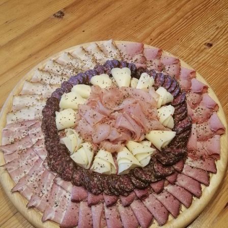 Ein Holzbrett zeigt eine kreisförmige Anordnung verschiedener Fleisch- und Käsesorten, darunter Schinken-, Salami- und Käsescheiben.