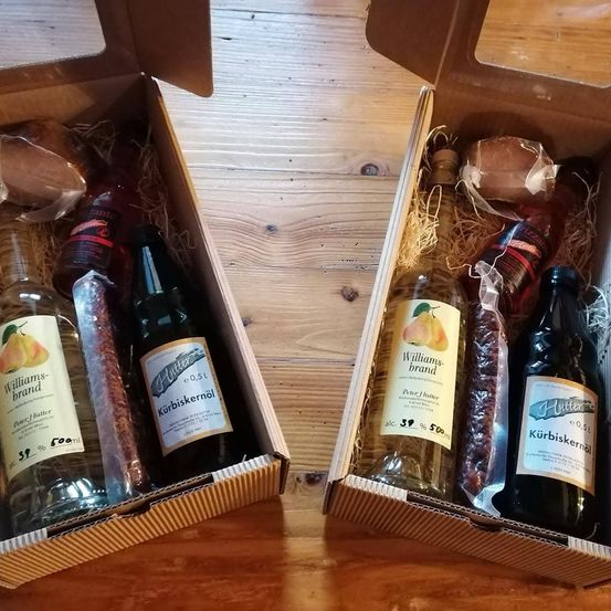 Zwei offene Kartons auf einem Holztisch, jeder mit einer Auswahl an Flaschen und verpackten Gegenständen. Ein Kasten enthält einen Williams-Birnenlikör und eine Flasche Kürbiskernöl, der andere hat einen ähnlichen Birnenlikör und eine weitere Flasche Kürbiskernöl.