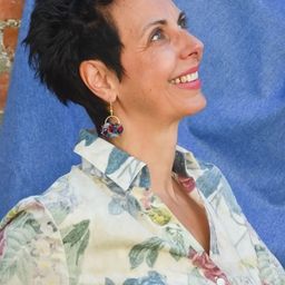 Eine Frau mit kurzen schwarzen Haaren lächelt und trägt ein Blumenhemd. Sie lehnt sich gegen einen blauen Stoffhintergrund.