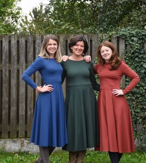 Drei Frauen stehen gemeinsam vor einem Holzzaun in einem Garten, jede trägt ein langärmeliges Kleid in verschiedenen Farben. Die Frau auf der linken Seite hat blonde Haare und trägt ein blaues Kleid. Die Frau in der Mitte mit kurzen Haaren trägt ein grünes Kleid, und die Frau auf der rechten Seite hat braune Haare und ein rotes Kleid.