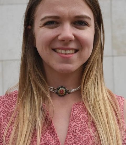 Eine Frau mit blonden Haaren, die ein rosa Blumenoberteil und eine Halskette mit einem rosenförmigen Anhänger trägt, lächelt und scheint für ein Foto zu posieren.