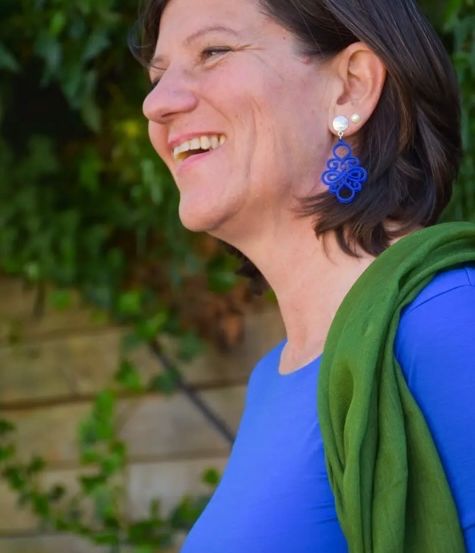 Eine Frau mit kurzen Haaren und blauen Ohrringen lächelt in die Kamera. Sie trägt ein blaues Hemd und einen grünen Schal. Hinter ihr ist eine Ziegelmauer mit viel Grün.