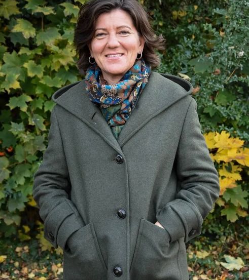 Eine Frau steht draußen in einem Waldgebiet, lächelt, trägt einen grünen Mantel und einen bunten Schal.