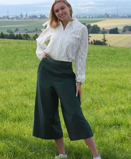 Eine Frau mit blondem Haar steht in einem grünen Feld, trägt eine weiße Bluse und grüne Hose. Sie posiert für ein Foto.