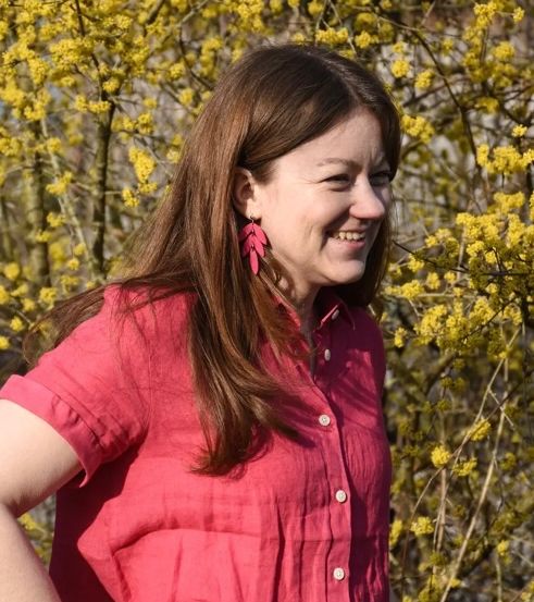 Eine Frau mit langen braunen Haaren, die ein rotes Hemd trägt, steht vor einem Busch mit gelben Blumen.