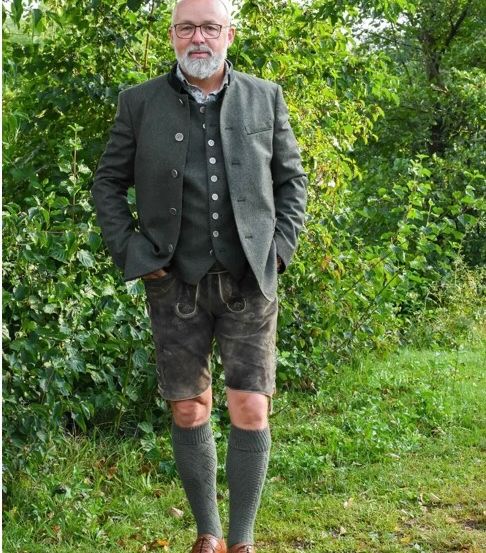 Ein älterer Mann mit Brille und Bart steht draußen auf einem Feld und trägt eine graue Jacke, Shorts und Socken.