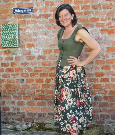 Eine Frau mit einem Blumenrock und grünem Oberteil steht lächelnd vor einer Ziegelmauer mit einem Schild, das Biergarten steht.
