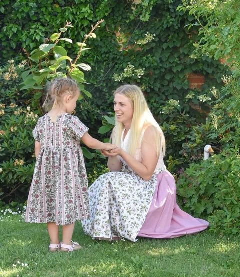 Eine Frau in einem Blumenkleid kniet auf dem Rasen, hält die Hand eines kleinen Mädchens und beide lächeln.