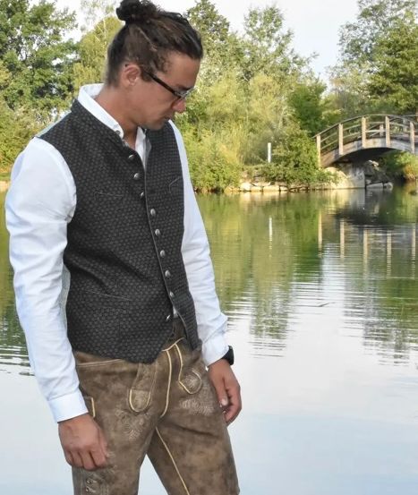 Ein Mann in traditioneller deutscher Kleidung steht am Ufer eines Sees und blickt in die Ferne. Er trägt eine schwarze Weste und braune Shorts. Hinter ihm überspannt eine Holzbrücke den See, und Bäume säumen das Ufer.