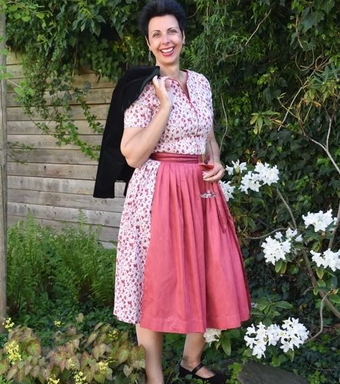 Eine lächelnde Frau steht im Garten, hält ein Weinglas in der Hand, trägt ein Blumenkleid und eine schwarze Jacke.