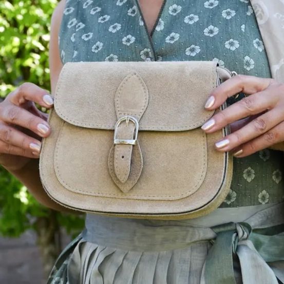 Eine Frau hält eine beige Wildleder-Umhängetasche mit einer silbernen Schnalle. Sie trägt ein Blumenkleid und hat weiße Nägel. Der Hintergrund zeigt Bäume.