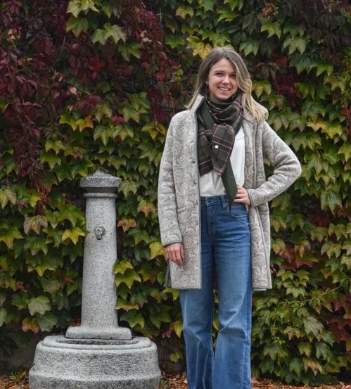 Eine Frau steht auf einem Bürgersteig vor einem steinernen Brunnen und trägt einen grauen Mantel, eine Schürze und Jeans. Sie lächelt mit den Händen auf den Hüften.
