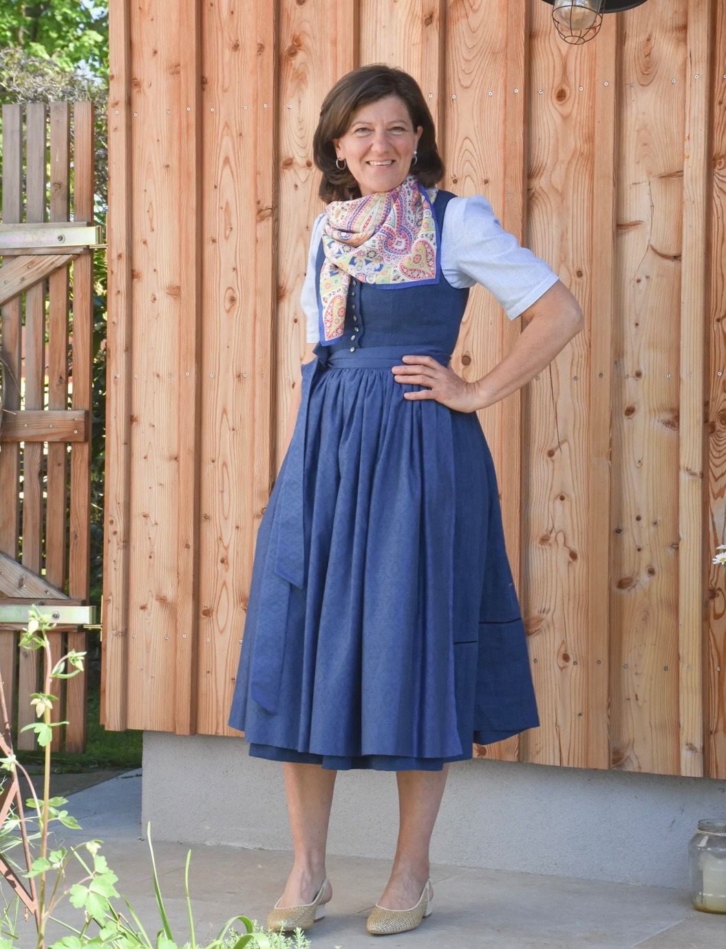 Eine Frau in einem blauen Kleid und einem Schal um den Hals steht vor einer Holzwand.